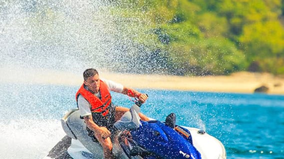 Rider wearing an orange life jacket on a jet ski, beach and palm trees in the background near Koh Samui